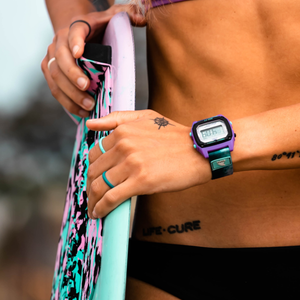 Person in a swimsuit holds a colorful surfboard, wearing Freestyle AMBER TORREALBA SIGNATURE SHARK CLASSIC CLIP REFLECTION watch.